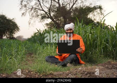 Indischer ländlicher Landwirt, der auf dem Feld der Feldfrüchte an einem Laptop arbeitet Stockfoto