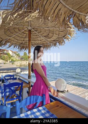 Ruhige Szene mit einer hübschen jungen Frau in rosa Kleid, die unter einem Vintage-Sonnenschirm auf einer Café-Lodge mit Blick auf die Küste steht und das B bewundert Stockfoto