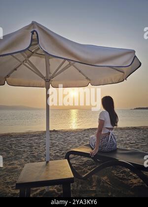 Charmante Frau saß auf einer Liege am Strand und beobachtete den Sonnenaufgang und nahm ein Sonnenbad am Morgen mit Blick auf das Meer. Wunderschöne Szenerie am Meer, s Stockfoto