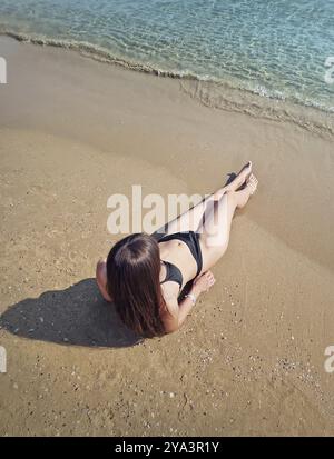 Charmante junge Frau im Bikini am Strand liegend und bräunend mit den Beinen zum Meer gestreckt. Klares Wasser schlägt sanft mit ihren Füßen. Ruhe und Entspannung Stockfoto