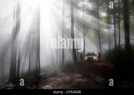 Paar steht neben einem Auto mit eingeschalteten Scheinwerfern, das auf einer nebligen Waldstraße geparkt ist, während dramatische Sonnenstrahlen bei Sonnenaufgang durch den Nebel scheinen. Stockfoto
