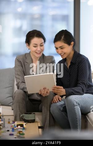 Zwei fröhliche, multiethnische Büroangestellte, die sich digitale Geräte teilen Stockfoto