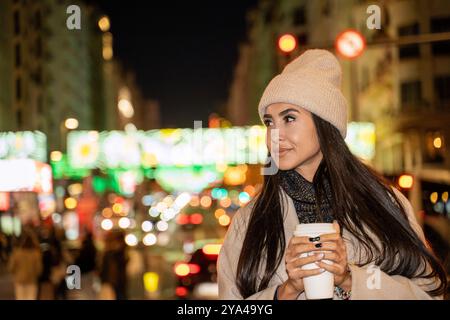 Eine glückliche Frau schlendert um Mitternacht durch die Stadt, genießt die Lichter der Stadt und hält eine Tasse Kaffee mit einem Lächeln im Gesicht Stockfoto