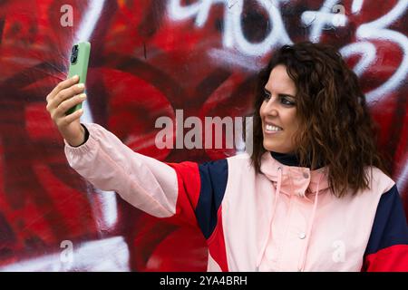 Eine junge Frau im Alter von 30 Jahren lächelt während eines Videogesprächs mit einer Wand, die mit roten Graffiti bedeckt ist und eine rosa Jacke trägt. Soziale Netzwerke Stockfoto