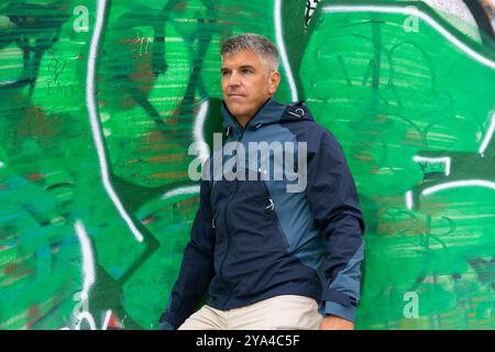 Grauhaariger Mann in seinen 40ern, der sich gegen eine leuchtend grüne Graffiti-Wand lehnt, einen blauen Regenmantel trägt und zur Seite blickt. Urbaner und legerer Street-Style Stockfoto