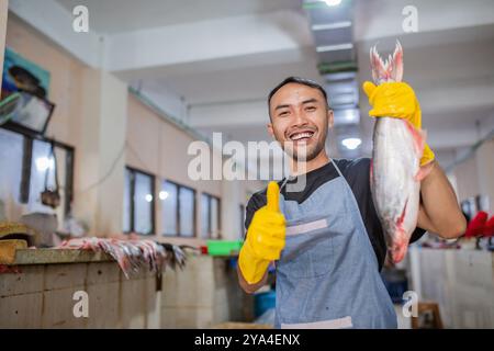 Fröhlicher Fischhändler, der fröhlich einen frischen Fang auf dem belebten Fischmarkt hält Stockfoto