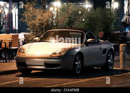 Ein silbernes Porsche Boxster Cabrio parkt vor einem beleuchteten Café in der Stadt und fängt die Eleganz und den Luxus eines Oldtimers in einem nächtlichen Ambiente ein Stockfoto