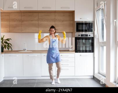 Fröhliche Frau in Schürze und gelben Schutzhandschuhen feiert eine saubere Küche nach der guten Arbeit des Housekeeping Stockfoto