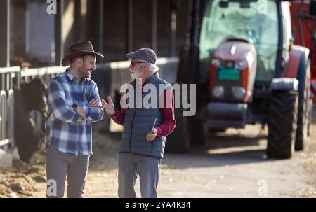 Zwei lächelnde Bauern reden vor Kuhstall und Traktor auf der Rinderfarm Stockfoto