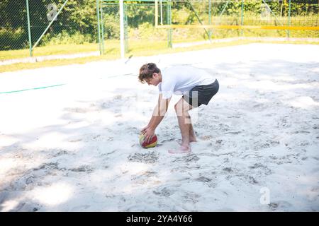 Der junge Mann beugt sich nach unten, um einen Volleyball vom Sandplatz abzuholen und bereitet sich auf das nächste Spiel vor. Die entspannte Umgebung im Freien ist von Grün umgeben, Stockfoto