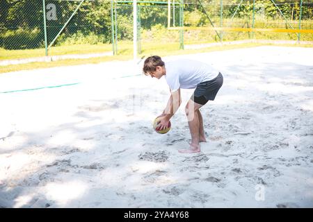 Der junge Mann beugt sich nach unten, um einen Volleyball vom Sandplatz abzuholen und bereitet sich auf das nächste Spiel vor. Die entspannte Umgebung im Freien ist von Grün umgeben, Stockfoto