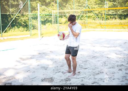 Der junge Mann beugt sich nach unten, um einen Volleyball vom Sandplatz abzuholen und bereitet sich auf das nächste Spiel vor. Die entspannte Umgebung im Freien ist von Grün umgeben, Stockfoto