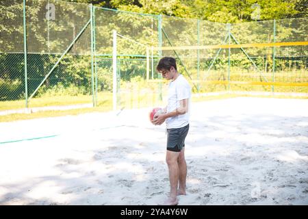 Der junge Mann beugt sich nach unten, um einen Volleyball vom Sandplatz abzuholen und bereitet sich auf das nächste Spiel vor. Die entspannte Umgebung im Freien ist von Grün umgeben, Stockfoto