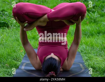 Junge braunhaarige Frau, die eine invertierte Yoga-Pose auf ihrer Matte im Freien in der Natur in einem schönen roten Outfit, perfekt für Yoga, spielt Stockfoto