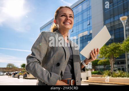 Moderne kaukasische Unternehmerin steht vor dem Büro mit einem digitalen Tablet in der Hand und lächelnd. Unternehmensstil. Selbstbewusste weibliche Leadershi Stockfoto