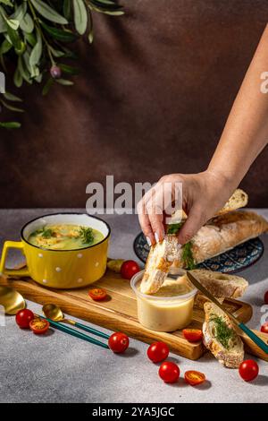 Eine Frau taucht ein Stück Baguette in Käseaufstrich, bereit zum Genuss mit einer Schüssel Brokkoli-Suppe Stockfoto