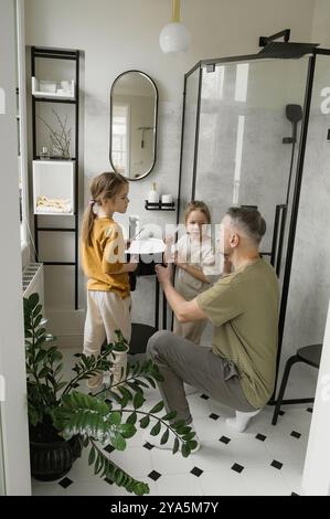 Ein Vater bringt seinen beiden Töchtern bei, wie man sich in einem modernen Badezimmer die Zähne putzt. Die Mädchen sehen ihren Vater an, während er ihnen zeigt, wie man putzt. Stockfoto