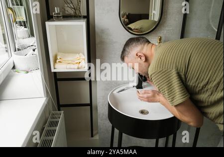 Ein Mann wäscht sein Gesicht am Waschbecken in einem stilvollen Badezimmer. Das Badezimmer ist mit weißen Wänden, schwarzen Akzenten und einem eleganten, modernen Design ausgestattet. Stockfoto