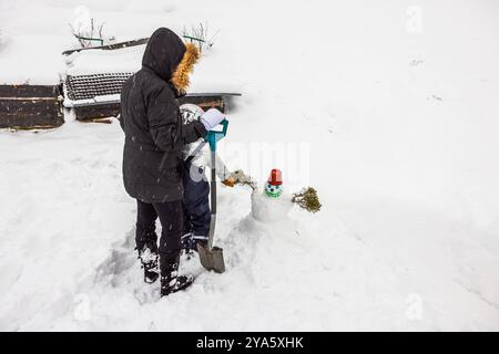 Mutter und Kind machen Schneemann mit rotem Eimer auf dem Kopf während des Schneefalls im Hinterhof der Villa. Schweden. Stockfoto