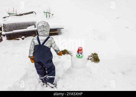 Kind im Winter, das einen kleinen Schneemann mit Topf für Hut im Hinterhof der schneebedeckten Villa baut. Schweden. Stockfoto