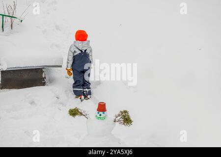 Kind in Winterkleidung steht im Schnee in der Nähe eines kleinen Schneemanns mit rotem Topf auf dem Kopf. Schweden. Stockfoto