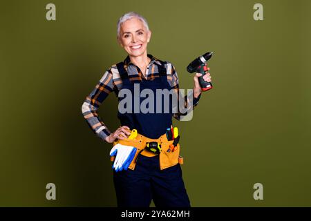 Foto-Porträt der attraktiven Reifen Frau professionelle Mechaniker halten Bohrer gekleidet blaue Arbeitskleidung insgesamt isoliert auf khakifarbenem Hintergrund Stockfoto