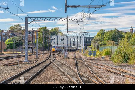 Ein Zug fährt von einem Bahnhof ab und unterquert eine eiserne Brücke. Eisenbahngleise treffen aufeinander und im Hintergrund befinden sich Häuser. Stockfoto