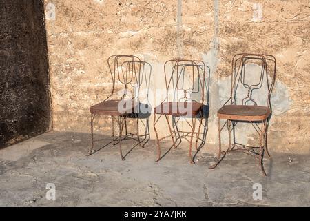 Drei alte, rostige Metallstühle stehen vor einer alten Wand in Indien als Symbol für die Zeit anh Geschichte und Leere Stockfoto