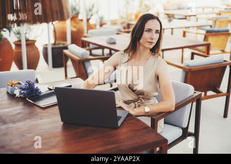Eine Geschäftsfrau sitzt mit Laptop und Notebook an einem hölzernen Tisch im Freien und trägt ein beigefarbenes ärmelloses Kleid. Blumen und Smartphone sichtbar. Hell und Stockfoto