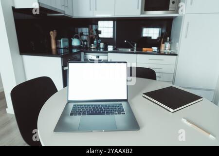 Eine saubere und moderne Küche mit weißen Schränken, Edelstahlgeräten und einer dunklen Arbeitsplatte. Ein Laptop mit leerem Bildschirm sitzt auf einem runden Tisch Stockfoto