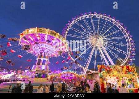 Ein lebhafter Jahrmarkt in der Abenddämmerung, beleuchtet von einem großen Riesenrad und einem bunten Kettenkarussell, Cannstadter Volksfest, Stuttgart-Bad Cannstadt, Baden-Wi¿½ Stockfoto