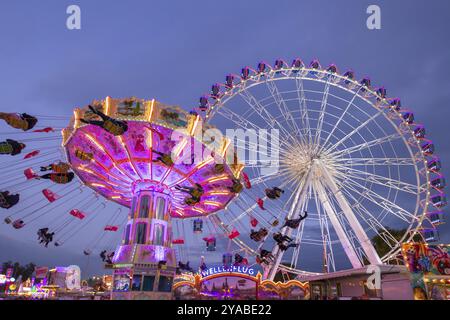 Ein lebhafter Jahrmarkt in der Abenddämmerung, beleuchtet von einem großen Riesenrad und einem bunten Kettenkarussell, Cannstadter Volksfest, Stuttgart-Bad Cannstadt, Baden-Wi¿½ Stockfoto