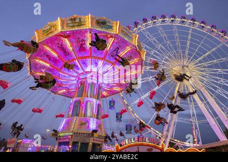 Ein lebhafter Jahrmarkt in der Abenddämmerung, beleuchtet von einem großen Riesenrad und einem bunten Kettenkarussell, Cannstadter Volksfest, Stuttgart-Bad Cannstadt, Baden-Wi¿½ Stockfoto