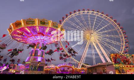 Ein lebhafter Jahrmarkt in der Abenddämmerung, beleuchtet von einem großen Riesenrad und einem bunten Kettenkarussell, Cannstadter Volksfest, Stuttgart-Bad Cannstadt, Baden-Wi¿½ Stockfoto