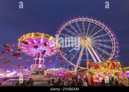 Ein lebhafter Jahrmarkt in der Abenddämmerung, beleuchtet von einem großen Riesenrad und einem bunten Kettenkarussell, Cannstadter Volksfest, Stuttgart-Bad Cannstadt, Baden-Wi¿½ Stockfoto