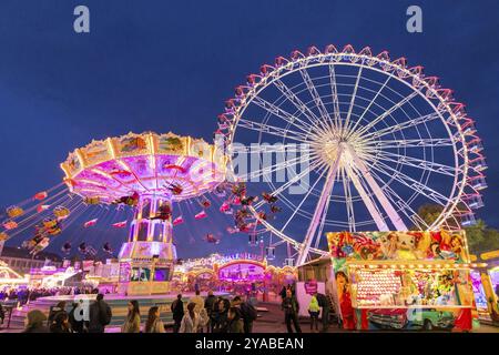 Ein lebhafter Jahrmarkt in der Abenddämmerung, beleuchtet von einem großen Riesenrad und einem bunten Kettenkarussell, Cannstadter Volksfest, Stuttgart-Bad Cannstadt, Baden-Wi¿½ Stockfoto