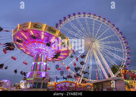 Ein lebhafter Jahrmarkt in der Abenddämmerung, beleuchtet von einem großen Riesenrad und einem bunten Kettenkarussell, Cannstadter Volksfest, Stuttgart-Bad Cannstadt, Baden-Wi¿½ Stockfoto