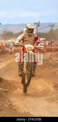 Motocross-Fahrer, der mit Schlamm und Staub bedeckt ist, springen mit seinem Motorrad über die Änderung der Steigung auf einer Feldstrecke während eines Motocross-Ausdauerrennens mit Zuschauern Stockfoto