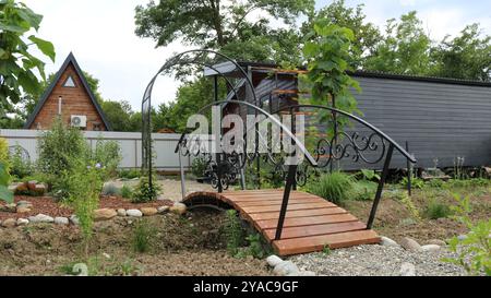 Kleine Bogenbrücke aus schwarzem Metall und braunem Holz in einem gepflegten Garten vor dem Hintergrund eines modernen einstöckigen hölzernen Cheroot-Hauses Stockfoto