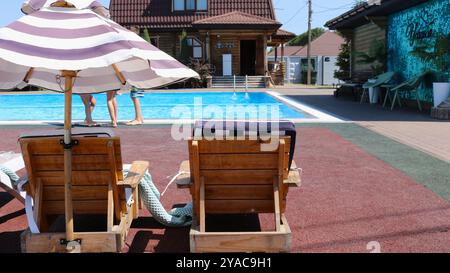 Neben einem Pool mit blauem, klarem Wasser stehen Holzliegen mit Sonnenschirmen vor dem Hintergrund eines zweistöckigen braunen Holzhauses Stockfoto