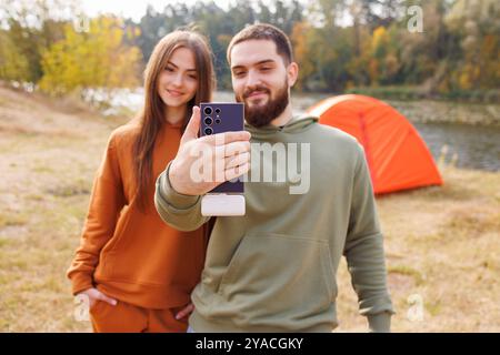 Ein junges Paar macht ein Selfie in einer malerischen Umgebung im Freien mit einem See und einem orangen Zelt im Hintergrund. Stockfoto