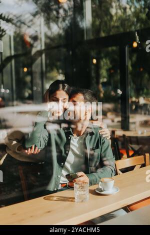 Junge Paare teilen sich zärtliche Momente in einem Café, umgeben von Grün und natürlichem Licht, während sie sich bei warmen Getränken und verspielten Gesten verbinden Stockfoto