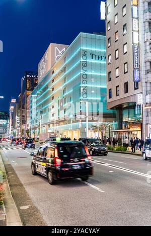 Entlang der Ginza mit dem Wahrzeichen Matsuya Ginza und dem Tiffany Store Gebäude beleuchtet in blauem, grünem, diffusem Licht. Im Vordergrund, Taxis fahren vorbei. Stockfoto