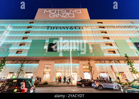 Auf der anderen Straßenseite bietet Ihnen ein flacher Blick auf das Wahrzeichen Matsuya Ginza und das Tiffany Store Building, beleuchtet in blauem, grünem Licht. Verkehr, der auf der Straße vorbeifährt. Stockfoto