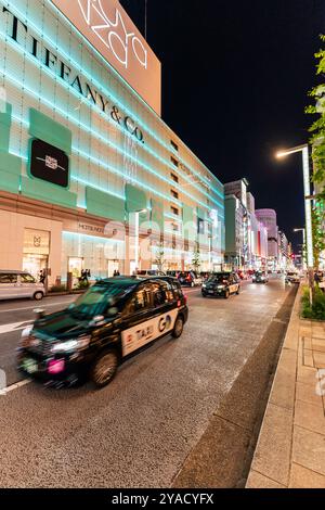 Mehrere Taxis fahren vorbei am Wahrzeichen Matsuya Ginza und dem Kaufhaus Tiffany, das nachts im grünen Licht vor dem schwarzen Nachthimmel beleuchtet wird Stockfoto