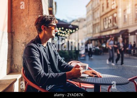 Porträt eines stilvollen Mannes, der im Café sitzt und Kaffee trinkt. Geschäftsmann genießt die Architektur der Stadt Lemberg auf der Straße Stockfoto