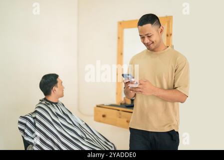 Ein Mann bekommt seinen Haarschnitt in einem Friseur, während ein anderer sein Handy überprüft Stockfoto