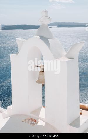 SANTORINI, GRIECHENLAND - 30. September 2024: Weißer Bogen mit Kirchenglocke mit blauem Wasser im Hintergrund. Stockfoto