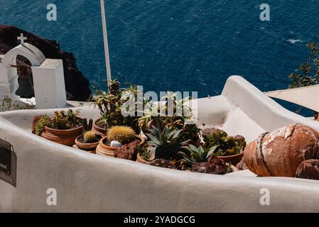 SANTORINI, GRIECHENLAND - 30. September 2024: Grüne Kakteen wachsen in braunen Tontöpfen auf der Terrasse eines weißen Hauses in Santorin mit Blick auf das ruhige Wasser Stockfoto