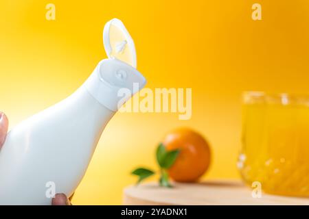 Nahaufnahme einer weißen Flasche mit einem Klappdeckel, teilweise geöffnet, auf einem hellgelben Hintergrund. Im Hintergrund steht ein Glas Orang Stockfoto